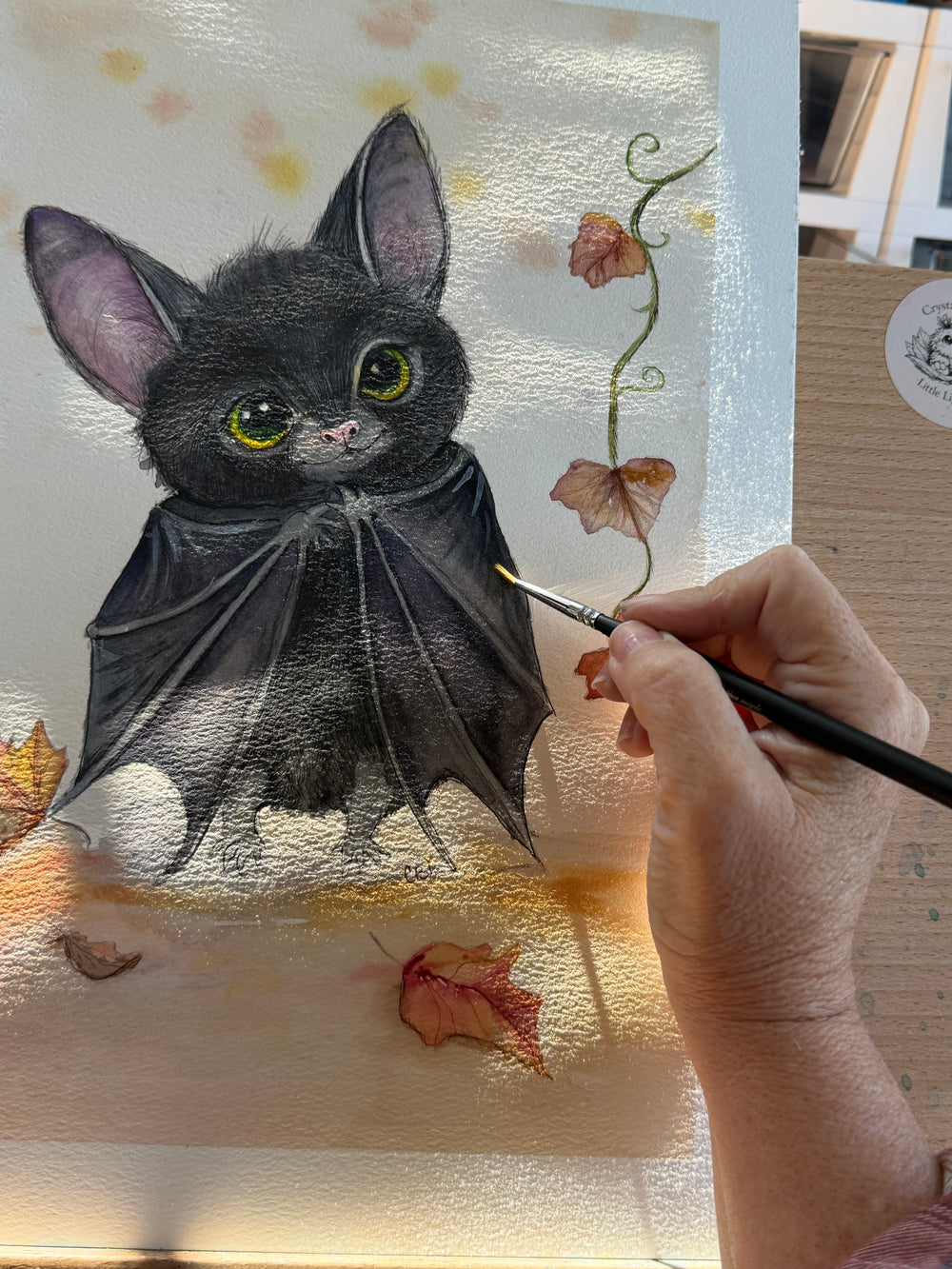 The artist’s hand holds a fine brush against a black bat watercolor painting surrounded by autumn leaves.