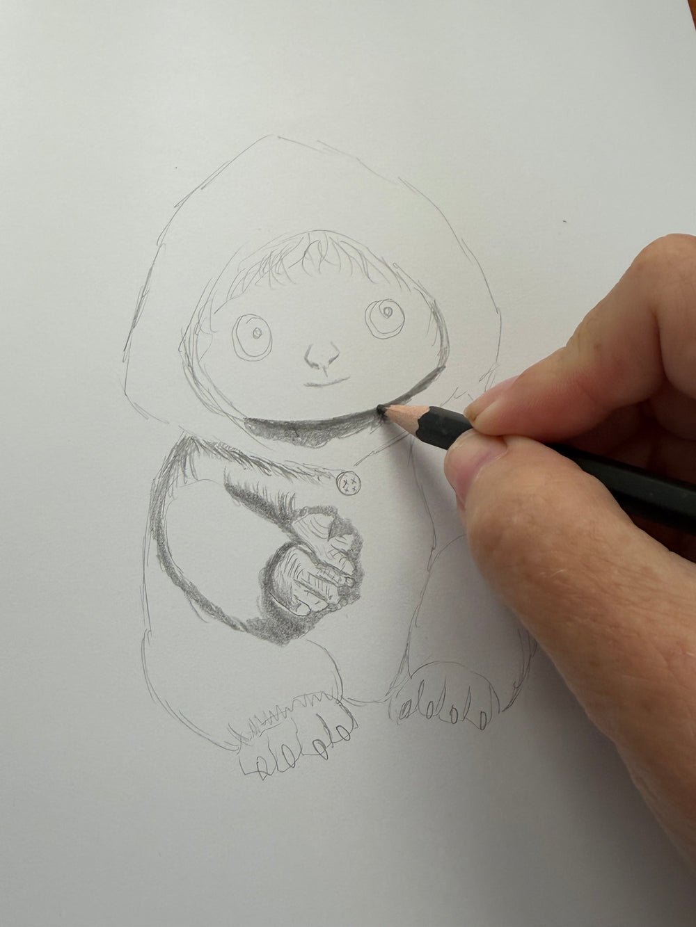 The artist’s hand shades a small hooded woodland creature in pencil on white paper.