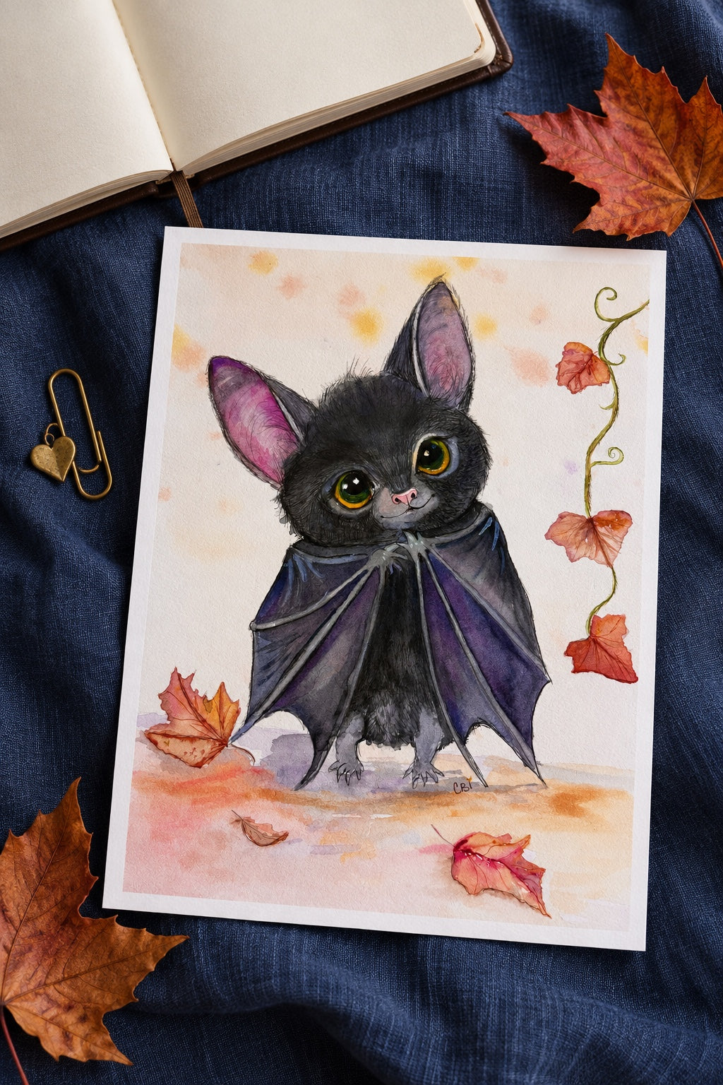 Unframed Batinsky print on blue linen beside an open blank notebook and autumn leaves.