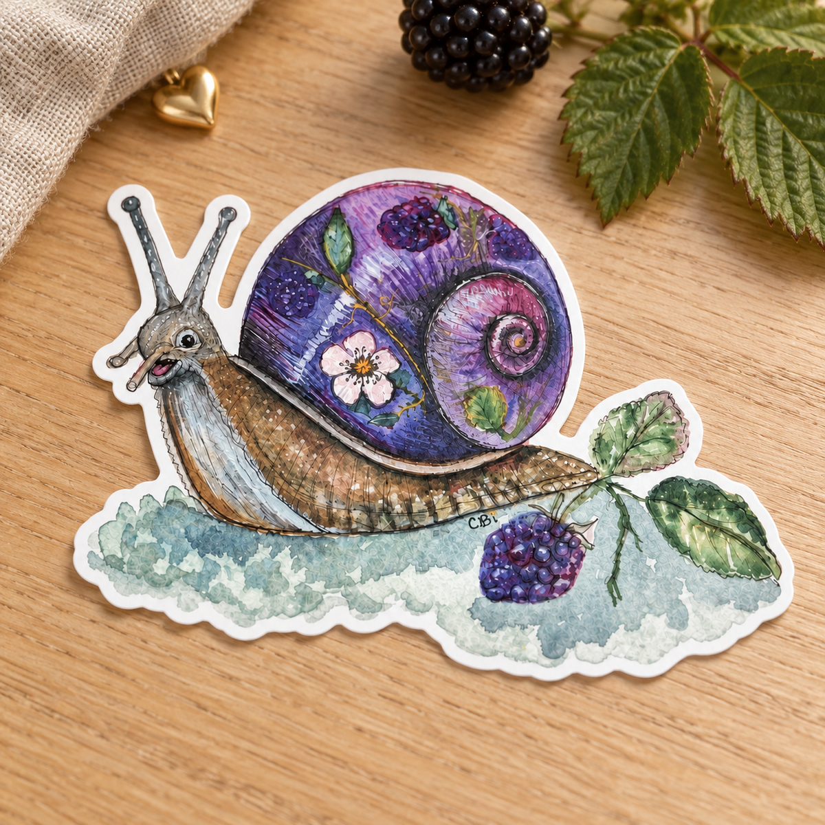 Bramble snail sticker photographed close up on oak with a blackberry, leaf, and linen.