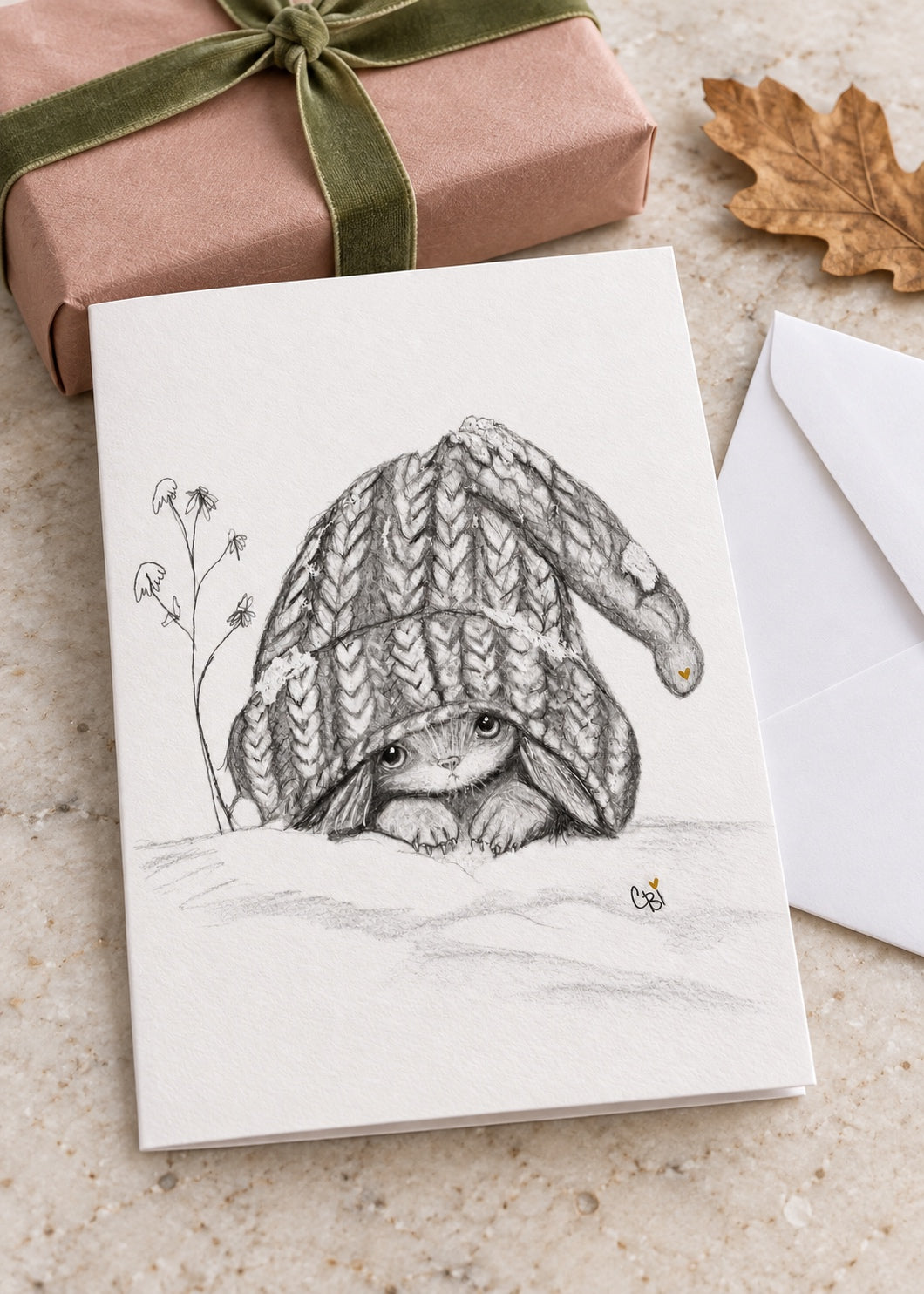 Caplings greeting card resting on a light stone table beside a rose-colored gift parcel, green ribbon, oak leaf and white envelope.