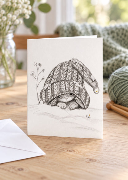 Caplings side-fold greeting card standing on a wooden knitting table beside sage yarn, knitting needles and a separate white envelope.