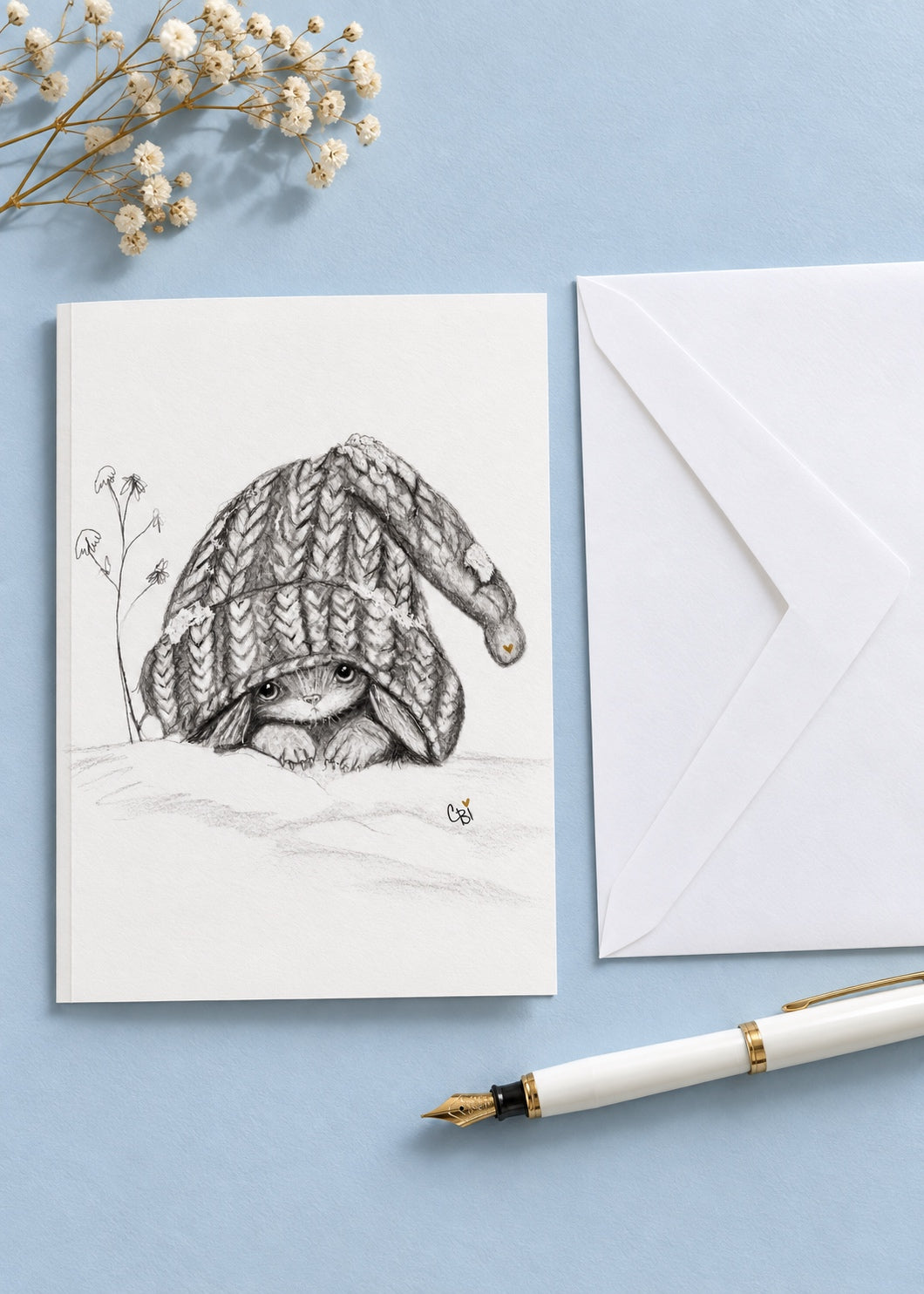 Caplings greeting card lying closed on a pale blue writing desk beside a white envelope, fountain pen and dried flowers.
