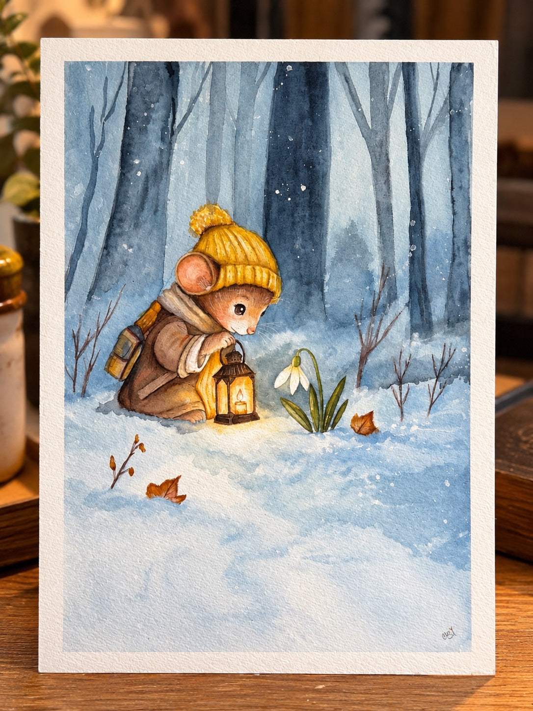 Cinder, a little mouse in a yellow knitted hat, holding a lantern beside a snowdrop in snowy blue woods; original watercolor photographed on a wooden table.