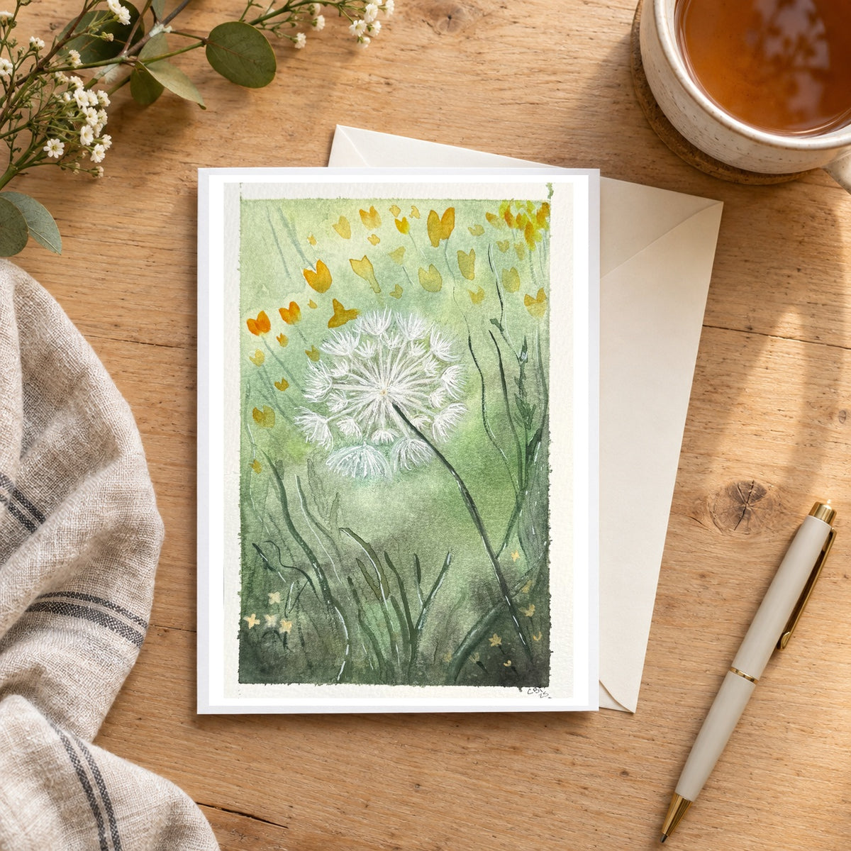 Dandelion greeting card with its original watercolor artwork and signature on a wooden desk beside tea, a cream envelope, a pen and flowers.