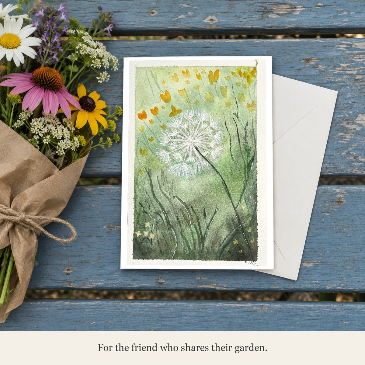 Dandelion greeting card beside garden flowers and an envelope on a blue bench. Caption: For the friend who shares their garden.