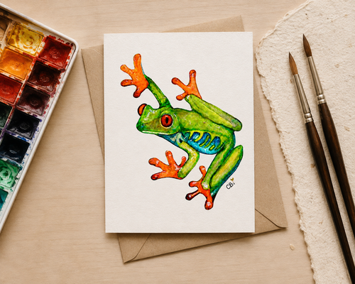 Hand holding a folded Red-Eyed Frog greeting card featuring a green watercolor frog with jewel-red eyes.