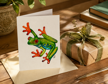 Folded Red-Eyed Frog greeting card beside a wrapped gift, showing a green watercolor frog among cattails and purple flowers.