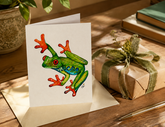 Folded Red-Eyed Frog greeting card beside a wrapped gift, showing a green watercolor frog among cattails and purple flowers.