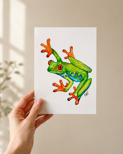 Red-Eyed Frog greeting card in an artist’s flat lay with the green watercolor frog and pond reflection visible.
