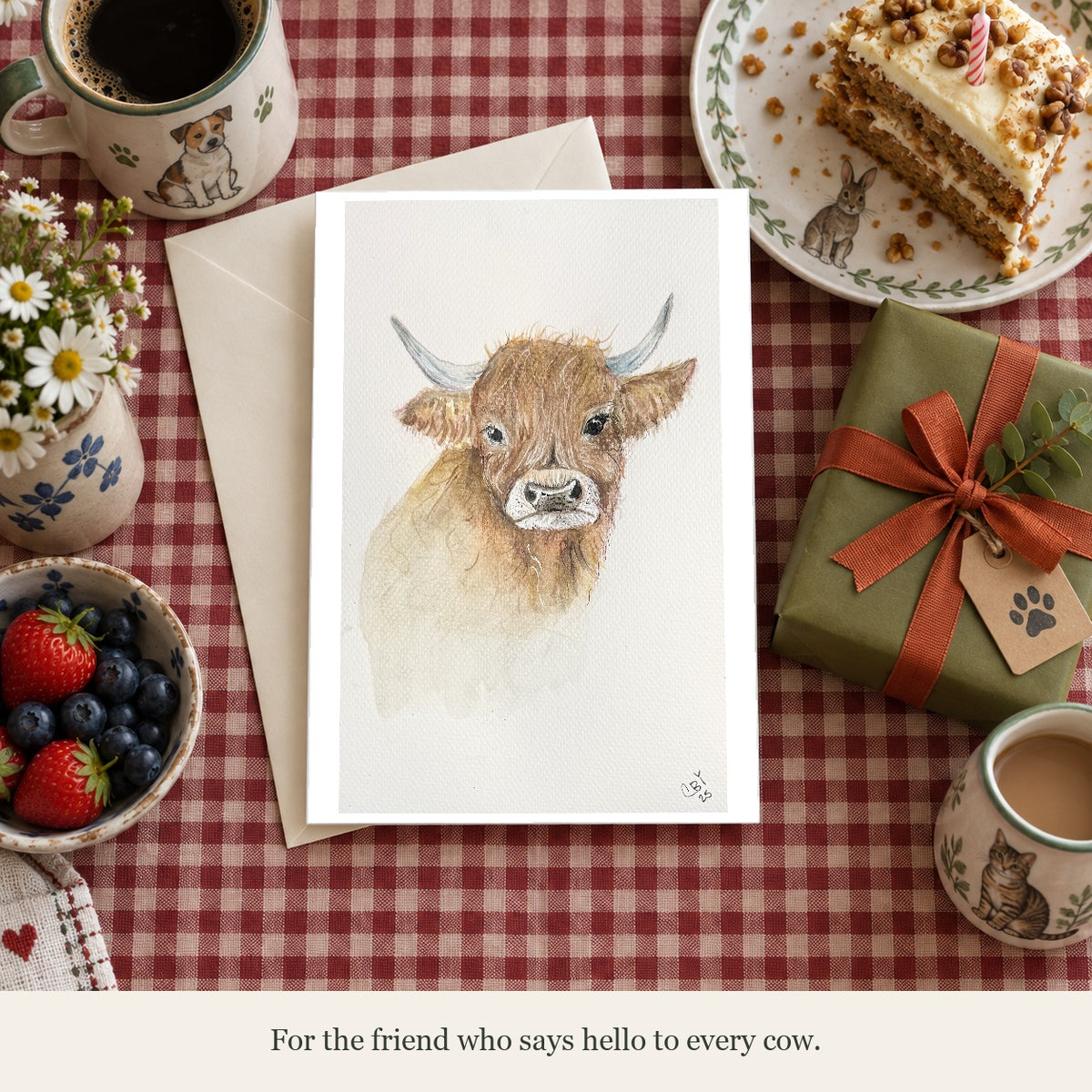 Highland cow greeting card beside a birthday gift and cake on a gingham table. Caption: For the friend who says hello to every cow.