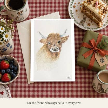 Highland cow greeting card beside a birthday gift and cake on a gingham table. Caption: For the friend who says hello to every cow.