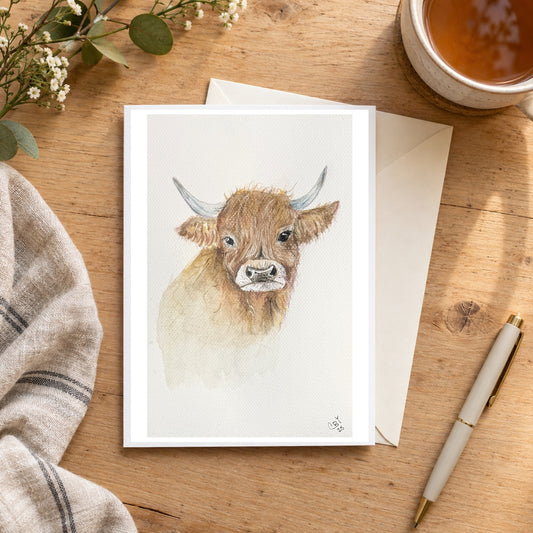 Highland cow greeting card on a wooden writing desk with tea, flowers, an envelope and pen.