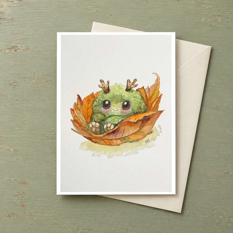 Fall Mossling greeting card featuring a green woodland creature nestled in autumn leaves, shown with an envelope.