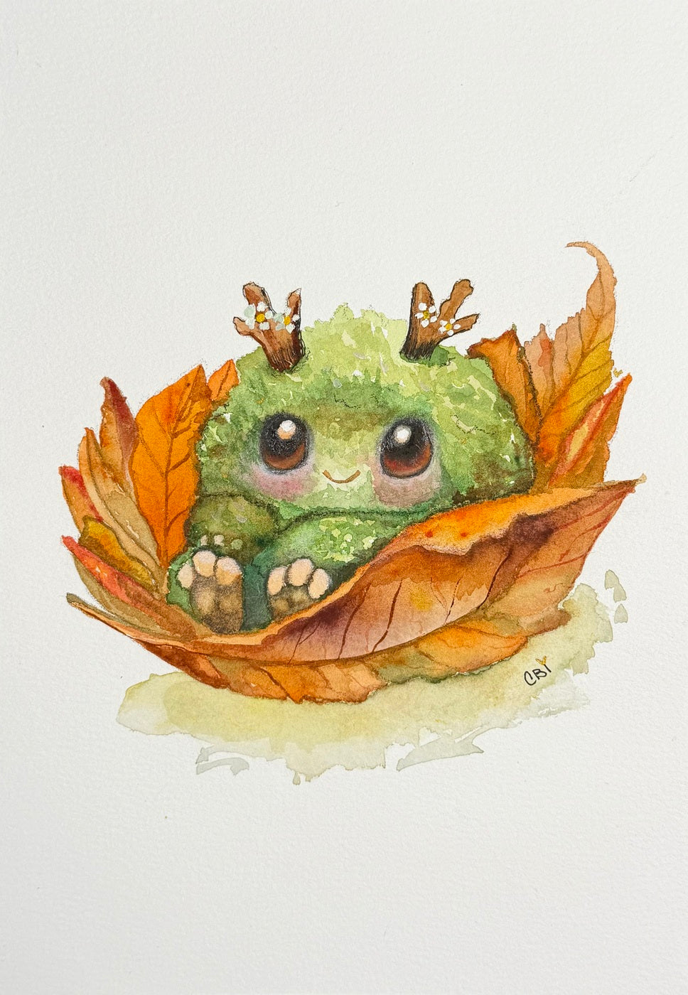 Complete original watercolor of a small green creature with twig antlers nestled in orange leaves, including the artist’s signature.
