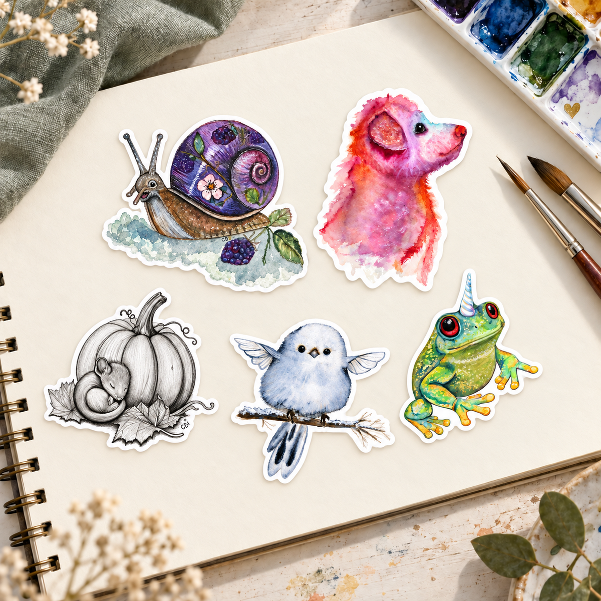 Five example vinyl stickers fully arranged on a cream sketchpad beside watercolor paints and brushes.