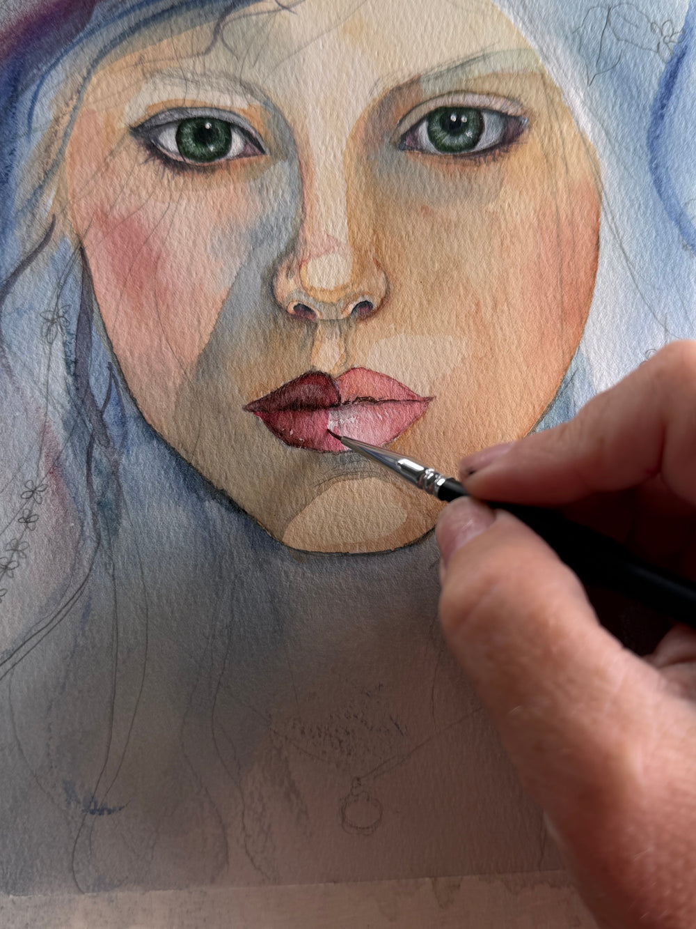 The artist’s hand uses a fine brush to paint the lips of a watercolor portrait.