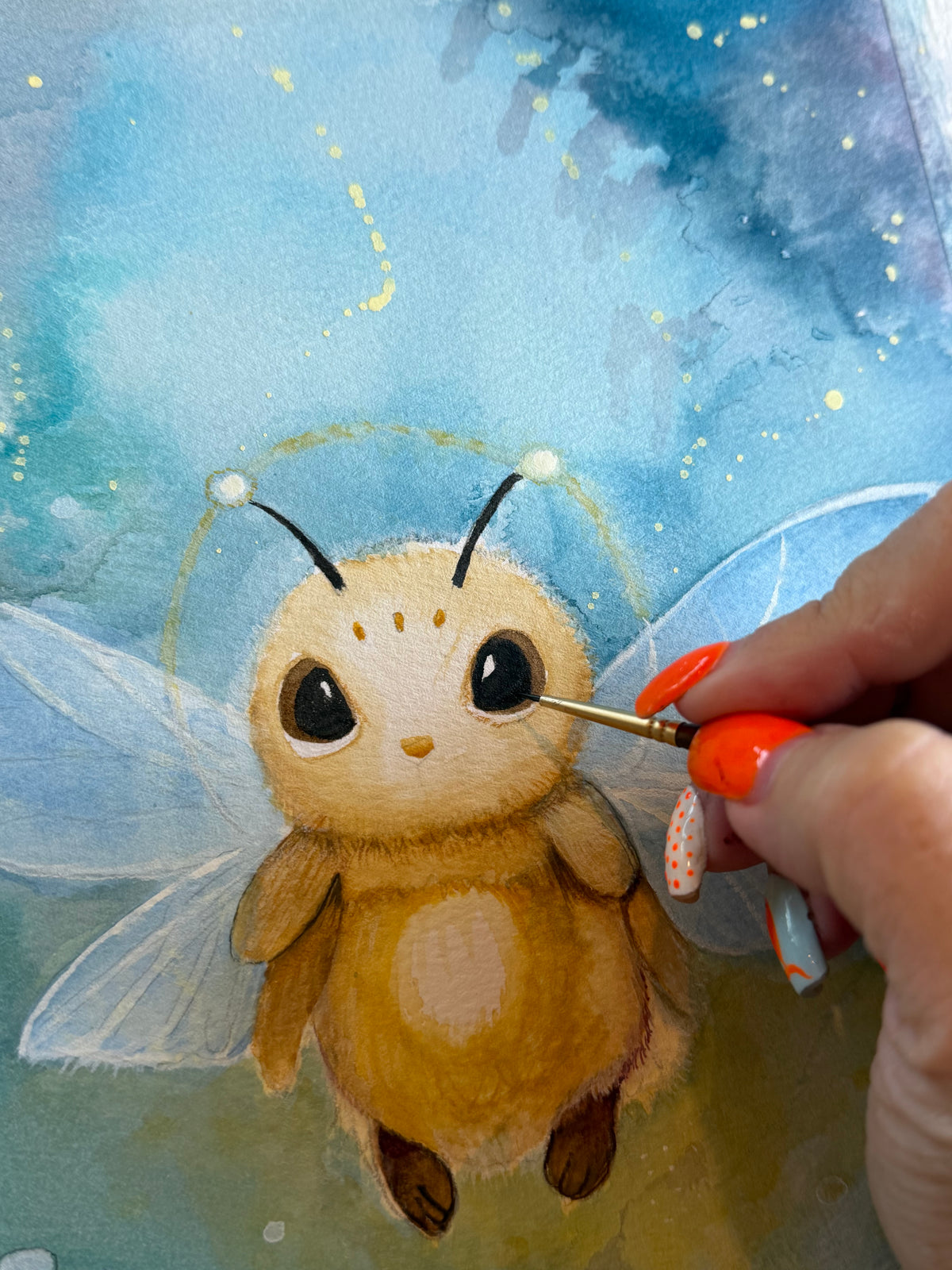 A hand using a fine brush to paint the eye of a golden winged creature on textured watercolor paper.