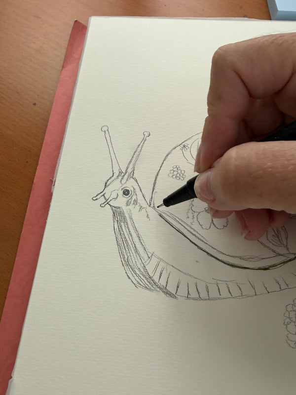 An artist’s hand drawing a snail with a patterned shell in pencil on paper.